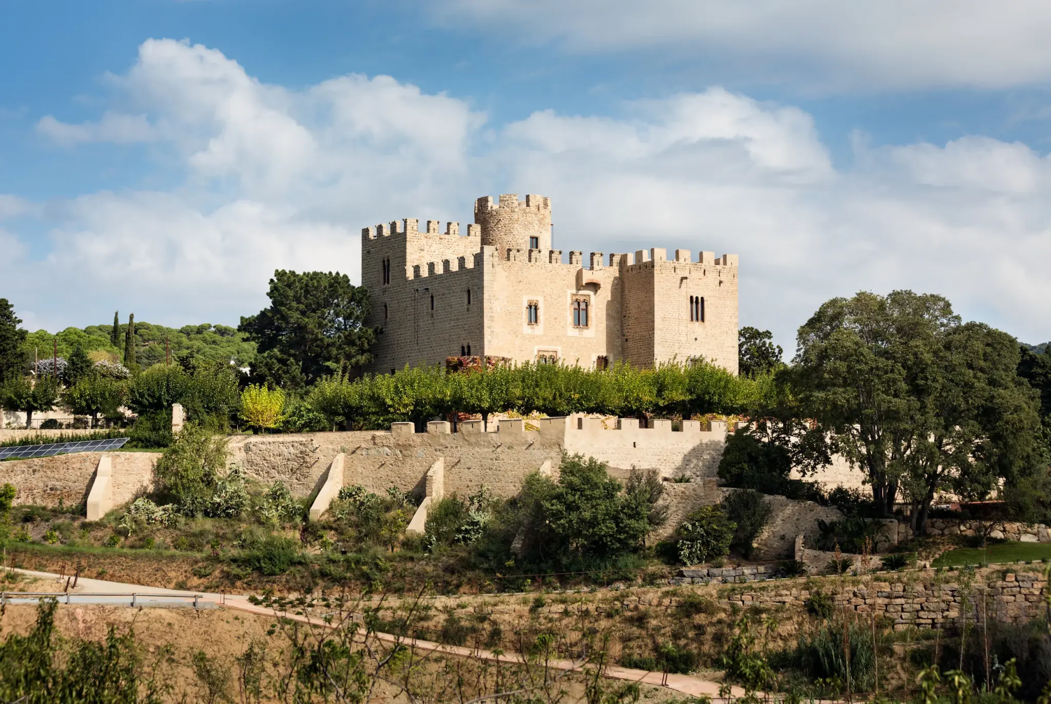 08-2048x1372 - Revela't 2026 Stone fortress with crenellated walls and a central tower, set among green trees under a blue sky with white clouds.