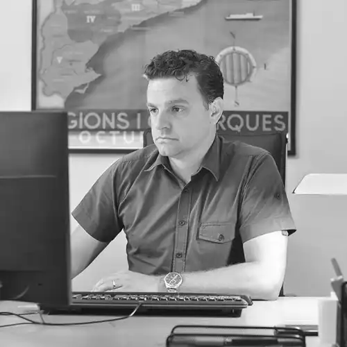 Man seated at a desk in an office, looking to the side, with a computer monitor and map poster in the background.
