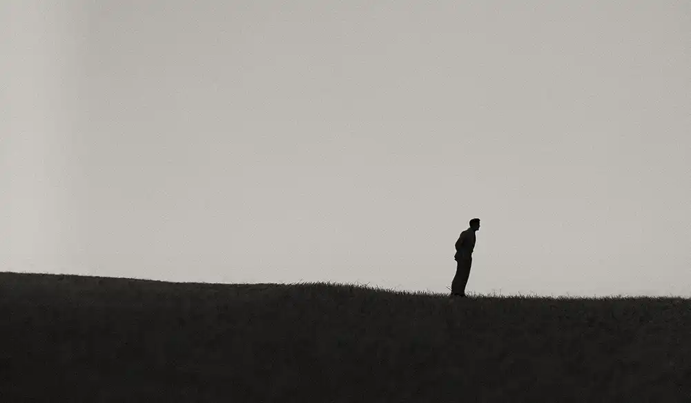 Silhouette of a lone man standing on a grassy hill against a pale sky.