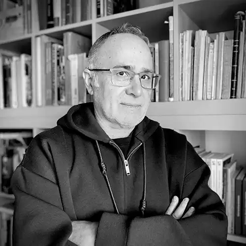 Portrait of a middle-aged man with glasses, wearing a dark hoodie, standing with arms crossed in front of a bookshelf in a grayscale photo.