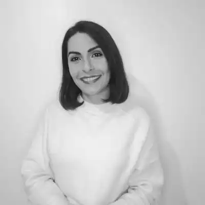 Portrait of a smiling woman with shoulder-length dark hair wearing a light sweater, standing against a plain wall.