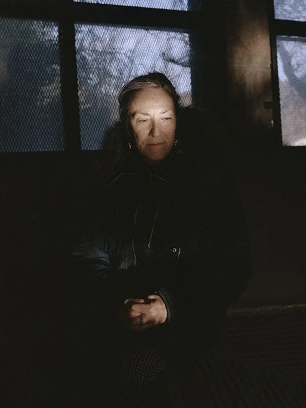 Older woman sits in a dim room with her eyes closed and hands folded in her lap.