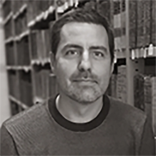 Portrait of a middle-aged man with short dark hair and light stubble, standing between tall shelves in a warehouse or storage area.