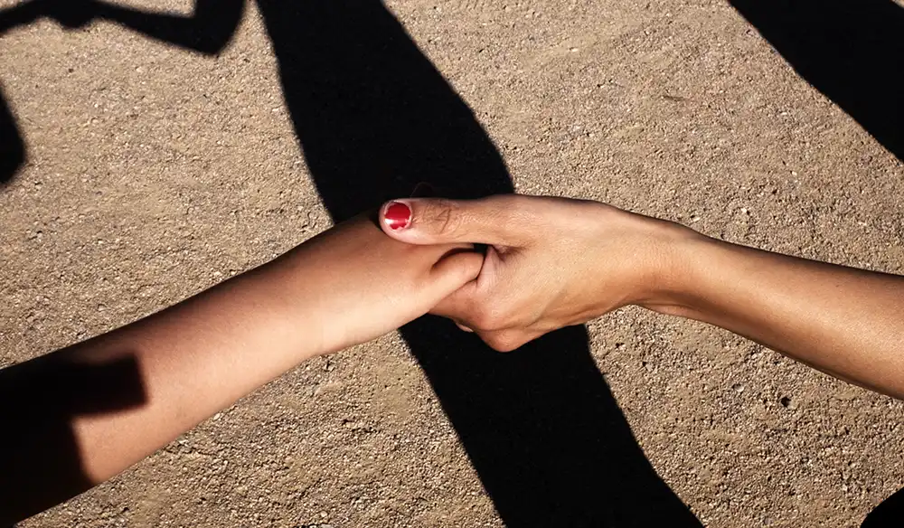 Imagen del taller el poble vertical - Revela't 2026 Two hands clasped in a handshake against a sandy ground with shadows nearby on the dirt surface.