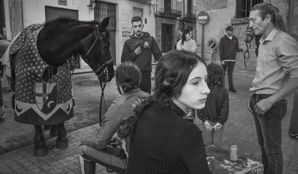 Young woman in the foreground on a cobblestone street; a patterned-blanket horse stands nearby as bystanders watch in the background.