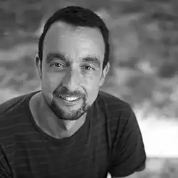 Portrait of a smiling man with short dark hair and a beard, wearing a dark striped shirt, outdoors (grayscale).