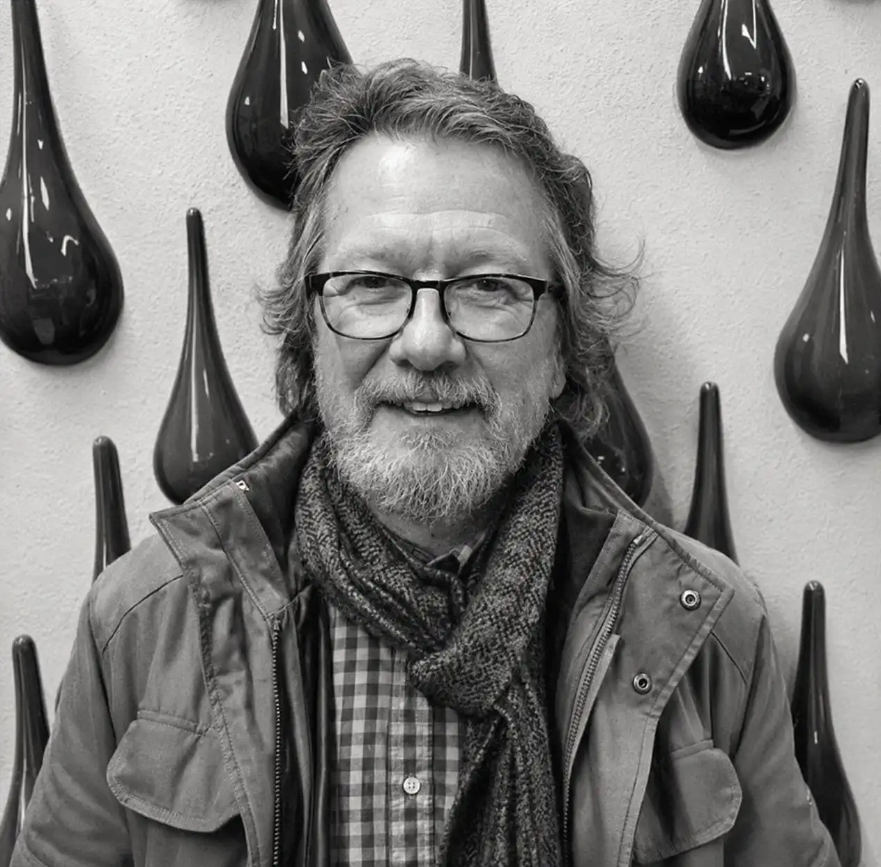 Portrait of a smiling man with glasses, a beard, scarf, and jacket, standing in front of a wall with glass drop sculptures (black-and-white photo).