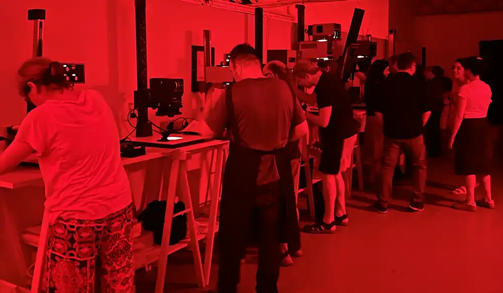 Laboratorio Revelat - Revela't 2026 People lined up at workstations with machines in a red-lit tech workshop inside a studio or lab.