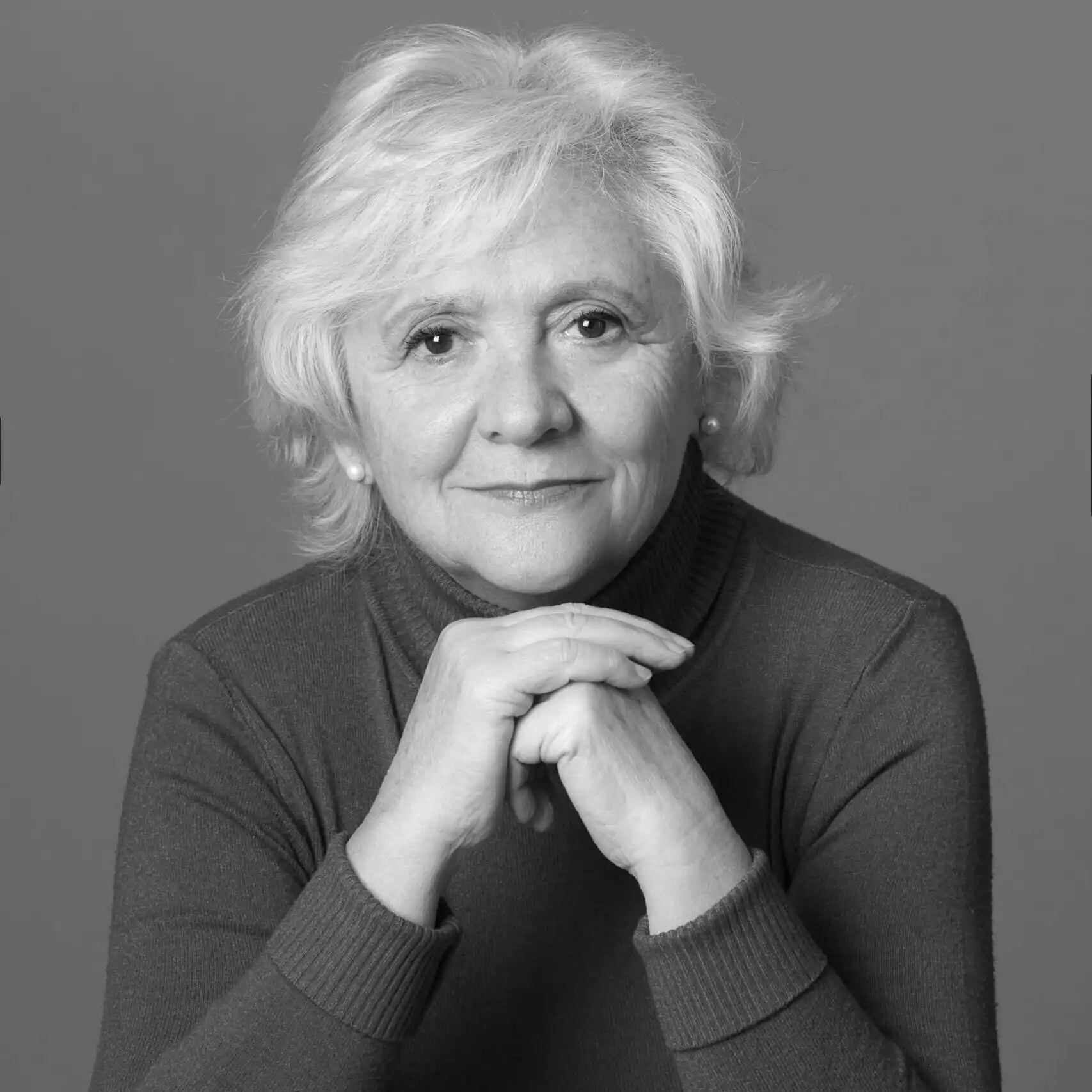 Portrait of an older woman with white hair resting her chin on clasped hands in a thoughtful pose.