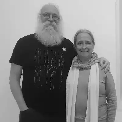A man with a long white beard and glasses stands with a smiling woman wearing a scarf indoors.