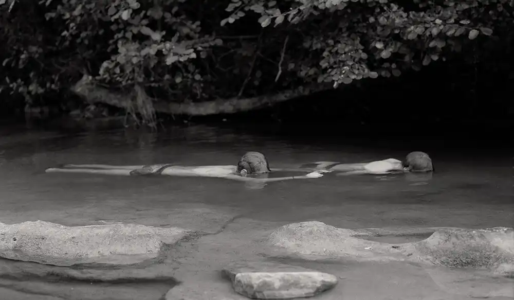 Black and white photo with two kids in the water, with trees on the background and stones in the front.