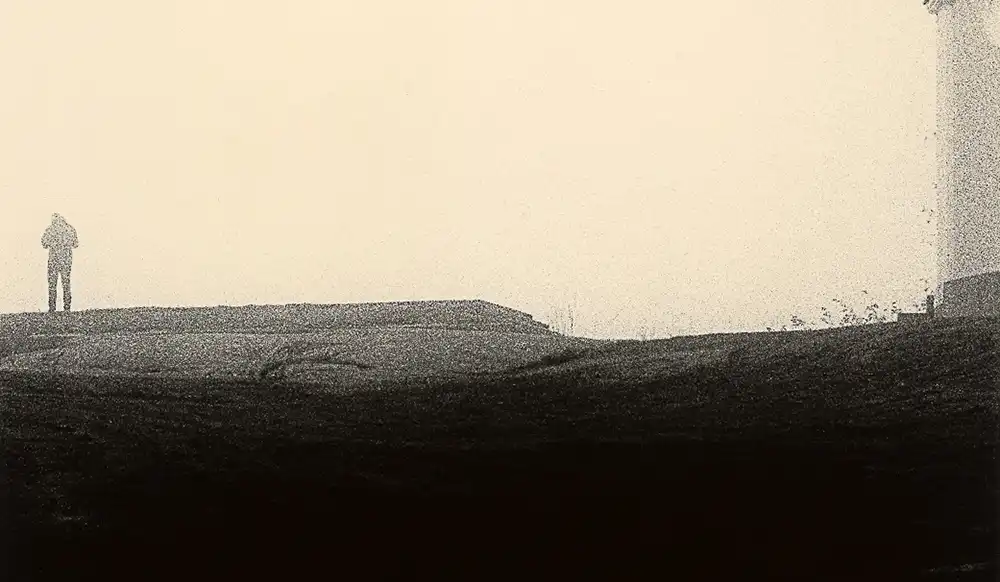 A lone figure stands on a hill at the edge of a desolate landscape under a pale sky.