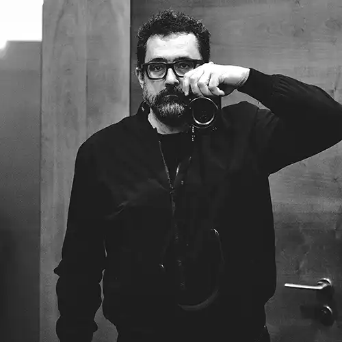 Bearded man wearing glasses and a dark sweater takes a mirror selfie with a camera held to his face.