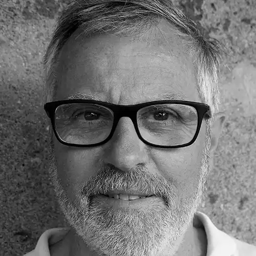 Close-up portrait of a middle-aged man with gray hair and beard, wearing black-framed glasses, looking at the camera against a textured wall.
