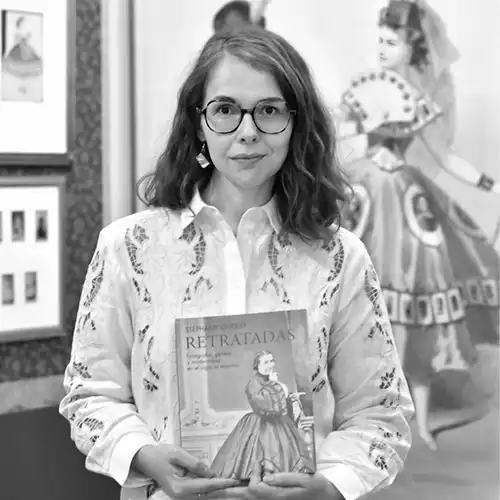 Woman with glasses and a white embroidered shirt holds a book titled 'Retratadas' in a museum or gallery setting, with art on the walls behind her.