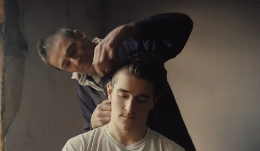 Man seated with his eyes closed as another man grips his neck from behind in a dimly lit room.guided by a tense moment