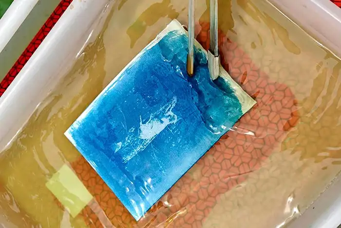 Blue dye swatch being dipped in a dye bath, held by tweezers over a tub with orange mesh pattern.