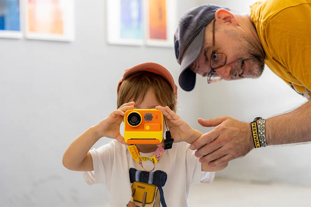 Child holds a bright yellow instant camera up to their face while an adult guides them nearby in a gallery-like setting.