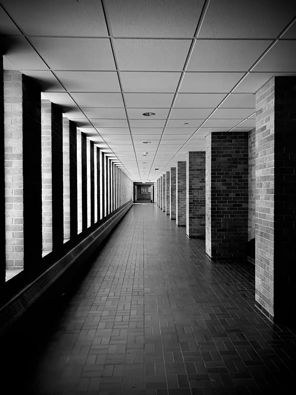 fugas - Revela't 2026 Black-and-white photo of a long, empty hallway with brick columns along both walls and a tiled floor.