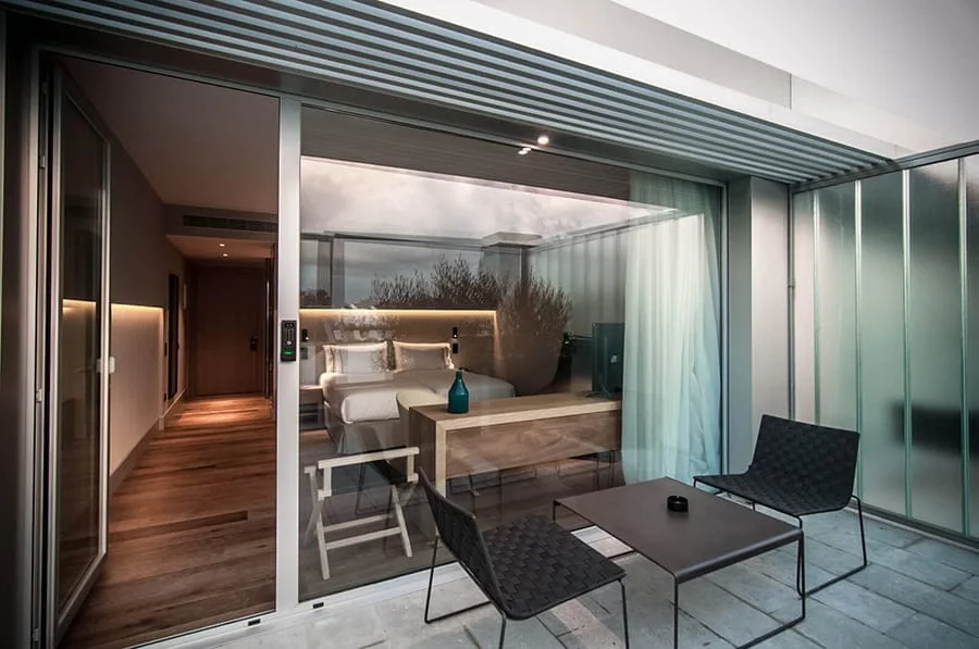 hotel_emocions - Revela't 2026 Modern hotel room seen through glass sliding doors; outdoor patio has two black chairs and a small table, bed and warm lighting inside.