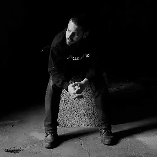 Black-and-white photo of a man sitting on a concrete block in a dark room, hands clasped, looking down.
