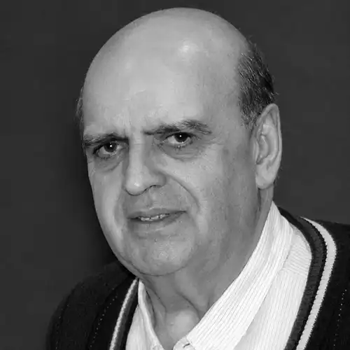 Older man with a bald head and light stubble wearing a white collared shirt and dark cardigan, looking at the camera in a grayscale portrait.