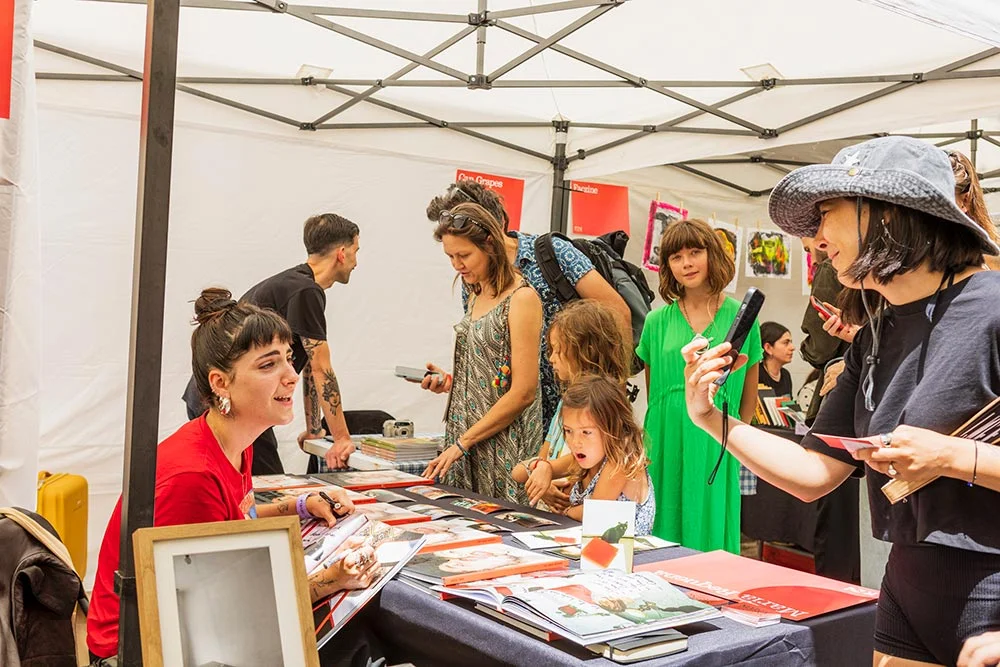 market2_ - Revela't 2026 Woman in red at a signing table under a tent, surrounded by visitors browsing books and chatting.