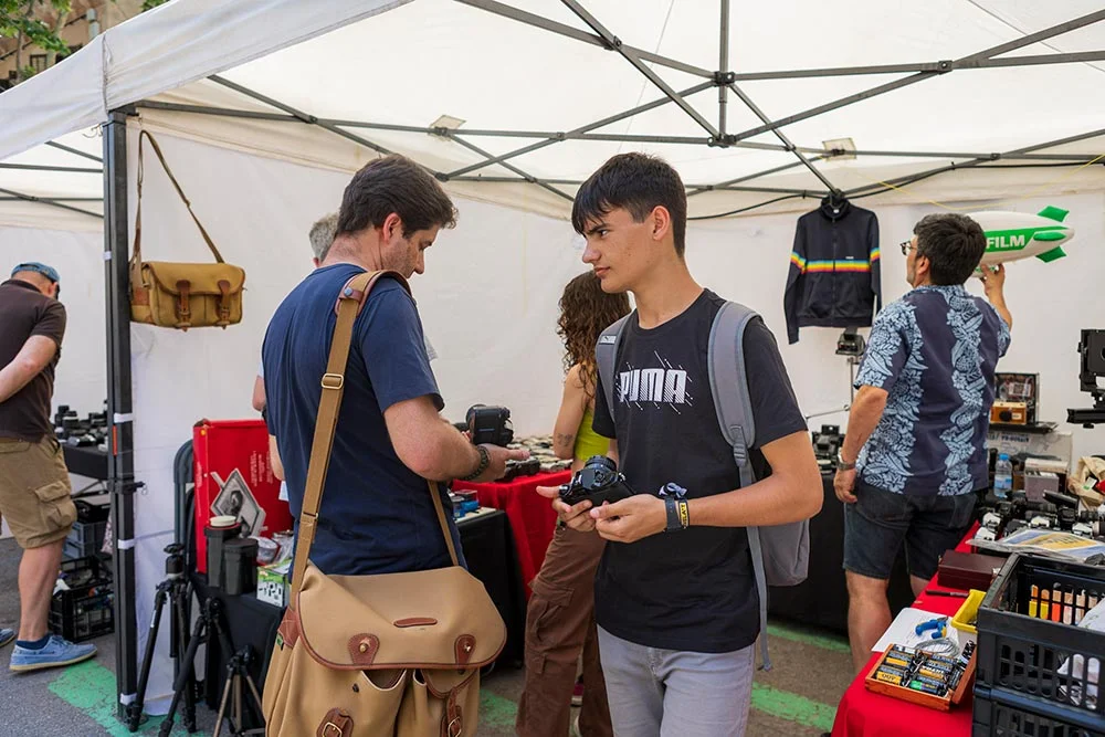 market3 - Revela't 2026 Group of young people at an outdoor gear market stall under a white canopy, browsing cameras and accessories on red tables.