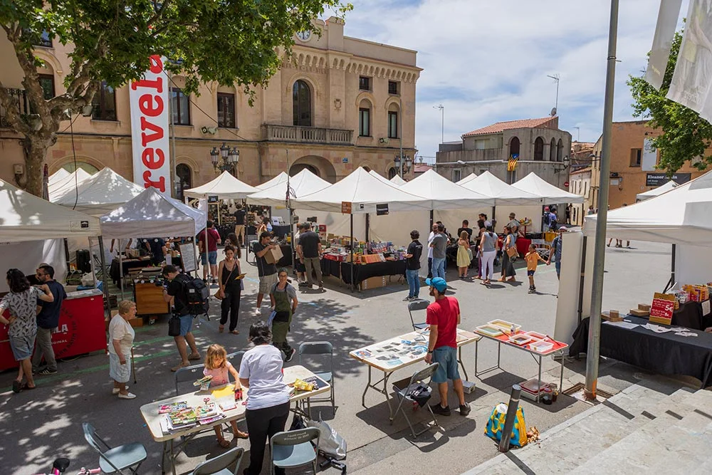 market4 - Revela't 2026 Outdoor marketplace with white tents and people browsing stalls in a town square on a sunny day