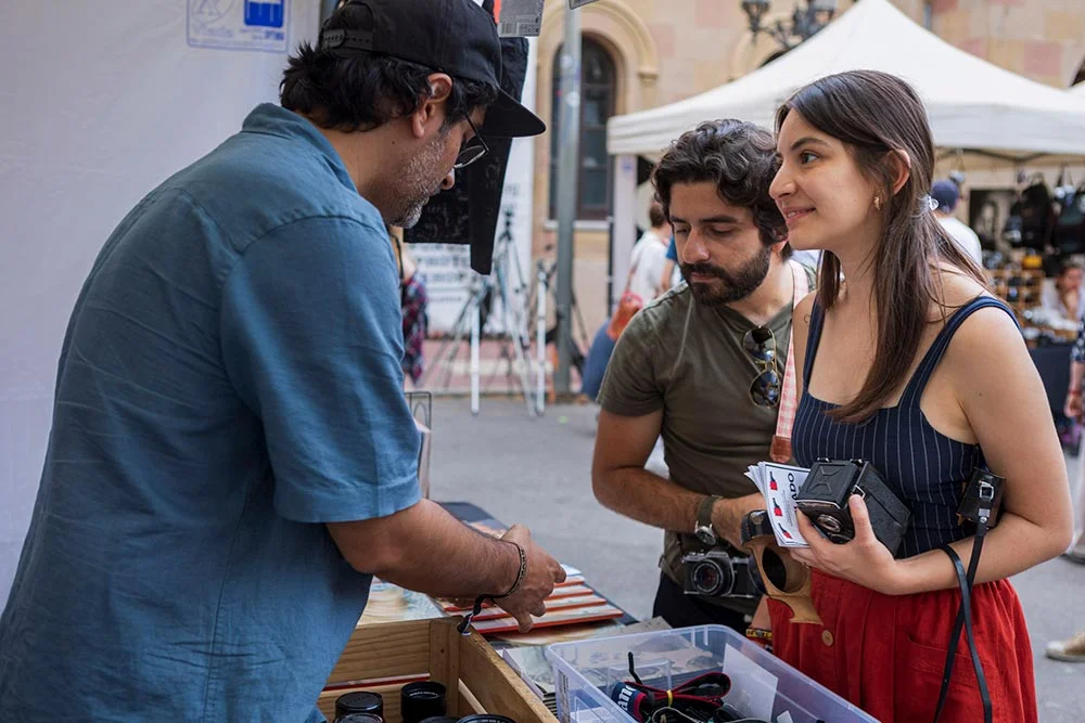market5 - Revela't 2026 Vendor at a street market sells items from a wooden stall to a smiling woman and a man who browse with cameras in hand.