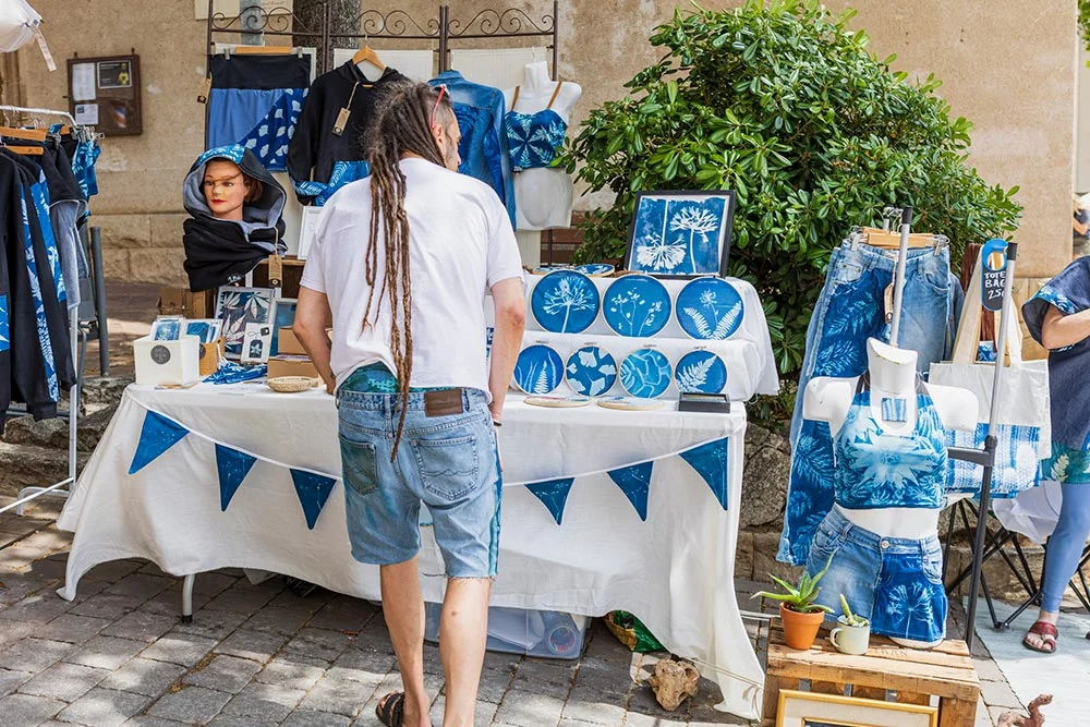 market6 - Revela't 2026 A man with long dreadlocks browses a blue-themed market stall displaying plates, clothing, and mannequins outdoors.