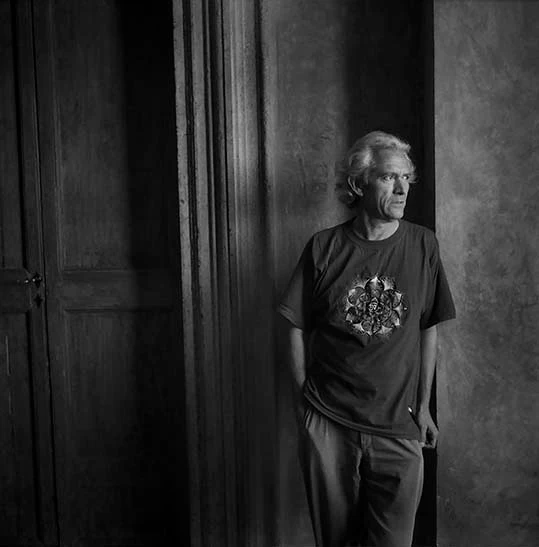 Older man with white hair stands in a doorway, wearing a dark graphic T-shirt and light pants, looking off to the side in moody lighting.