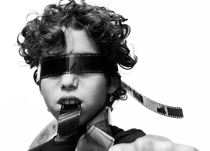 Black-and-white photo of a boy with film strips wrapped over his eyes and mouth, suggesting cinema or film.