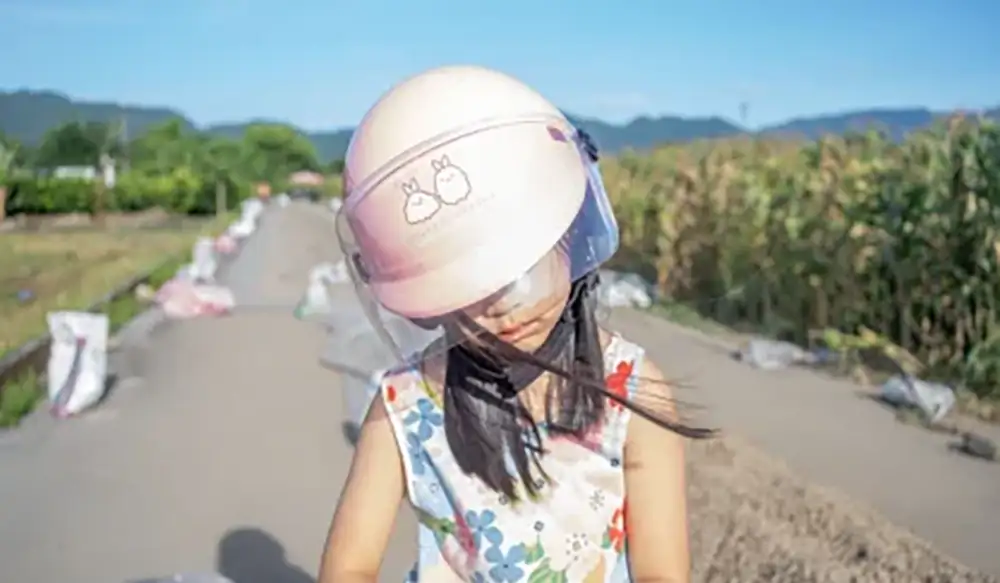 Young girl wearing a pink helmet with cartoon prints, standing on a rural road under construction with sand piles and green fields in the background.