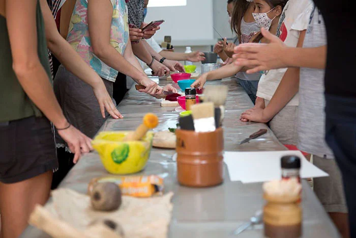 Group of people standing along a long table, reaching for colorful bowls and tools in a hands-on workshop (masks visible).