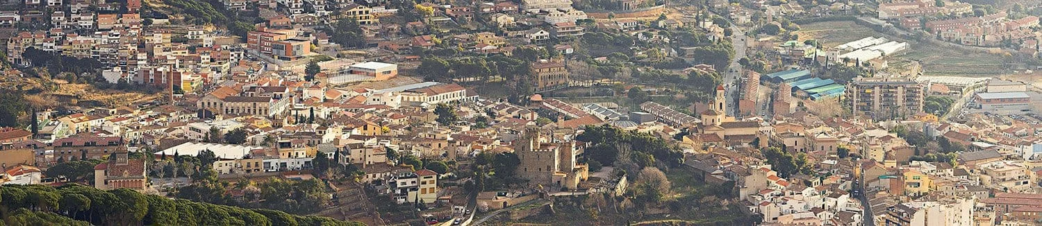 vilassar2 - Revela't 2026 Aerial view of a densely built hillside town with terracotta roofs, churches, and winding streets scattered among trees and hillsides.