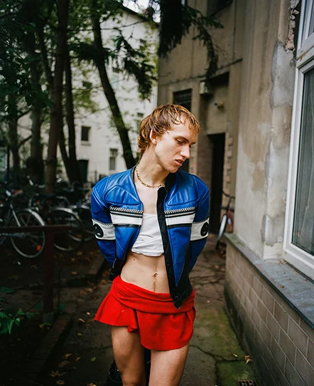 Young person in a blue cropped leather jacket, white crop top, and red mini skirt stands in an urban alley, looking down.