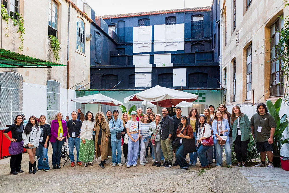 voluntariat - Revela't 2026 Diverse group of about 30 people posing for a group photo in a sunny courtyard with umbrellas.