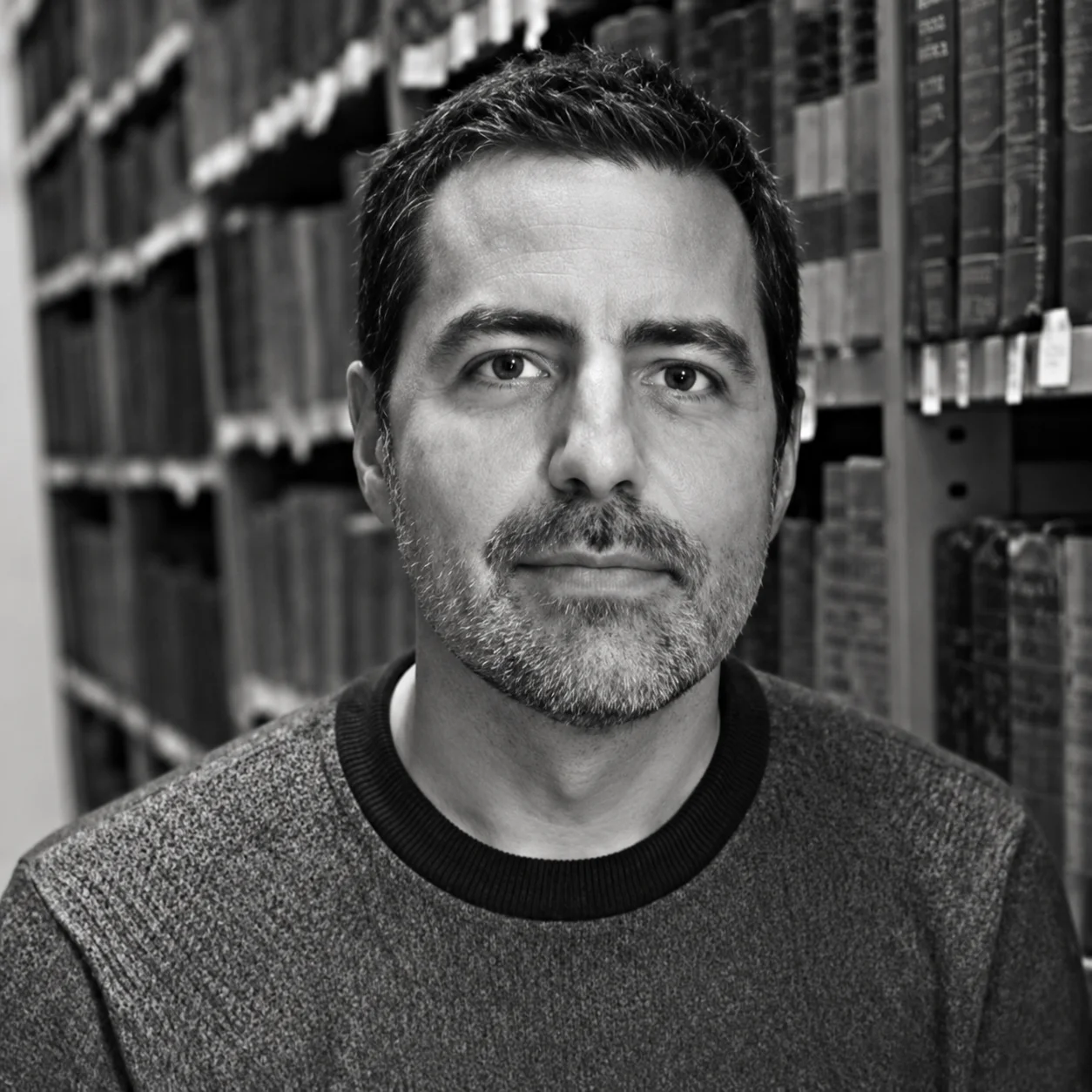 Black-and-white portrait of a man with short dark hair and stubble, standing in a library aisle with shelves behind him, looking at the camera.