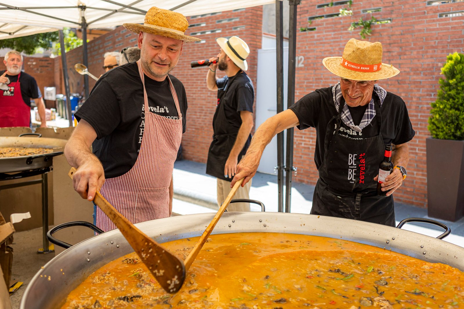 Revelat_25250525544 - Revela't 2026 Two men wearing straw hats and aprons stir a large paella pan of orange rice outdoors at a festival.