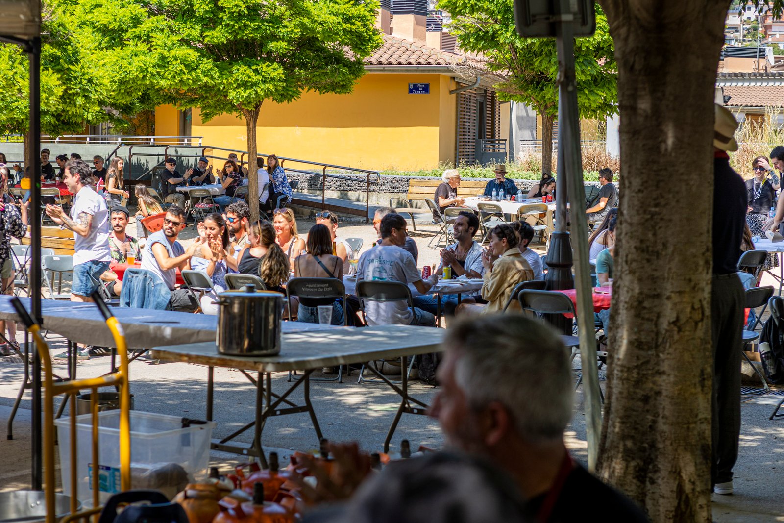 Revelat_25250525615 - Revela't 2026 Crowd of people sitting at outdoor tables under trees in a sunny plaza, enjoying drinks and conversation with a yellow building in the background