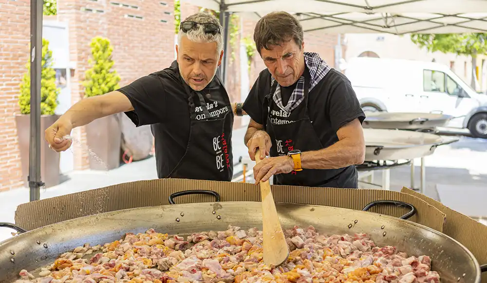 Paella Revela't 2025 - Revela't 2026 Two men wearing black shirts and aprons stir a massive paella pan filled with meat under a canopy outdoors.