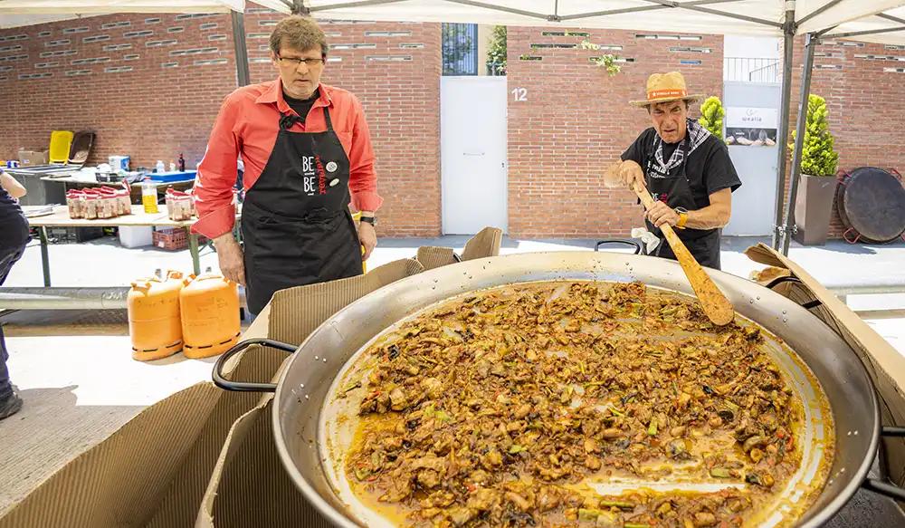 Paella Revela't 2025 - Revela't 2026 Two men cook a large paella-like dish in a wide pan under a canopy outdoors, with brick walls in the background.
