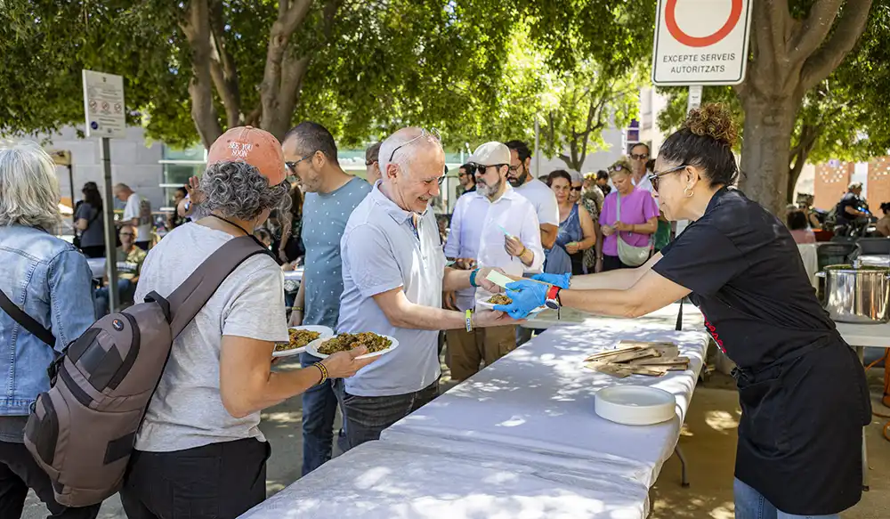 Paella Revela't 2025 - Revela't 2026 Volunteer wearing gloves hands a plate of food to a smiling man at an outdoor community meal under trees.