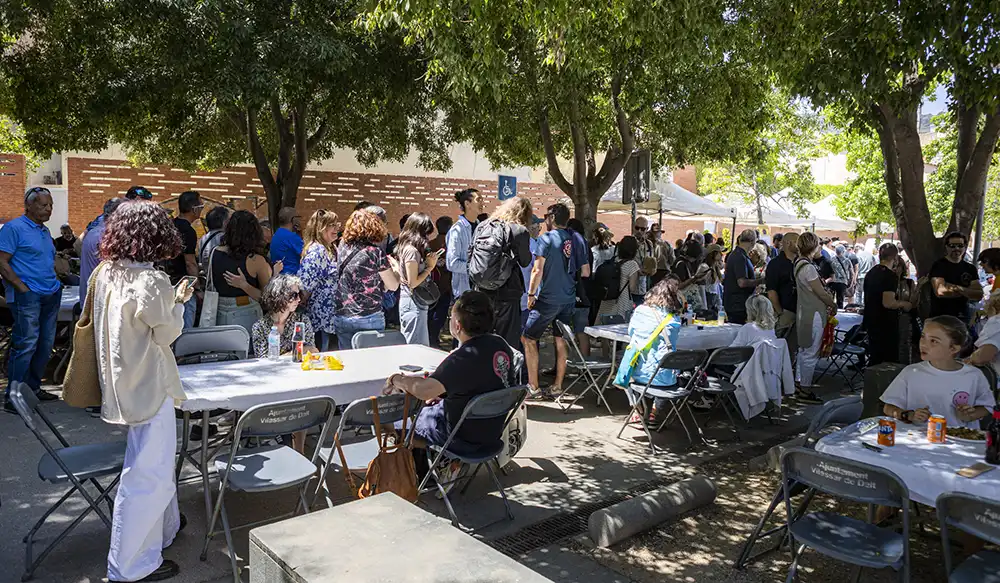 Paella Revela't 2025 - Revela't 2026 Crowd of people at an outdoor community event under trees with folding tables and tents in the background.
