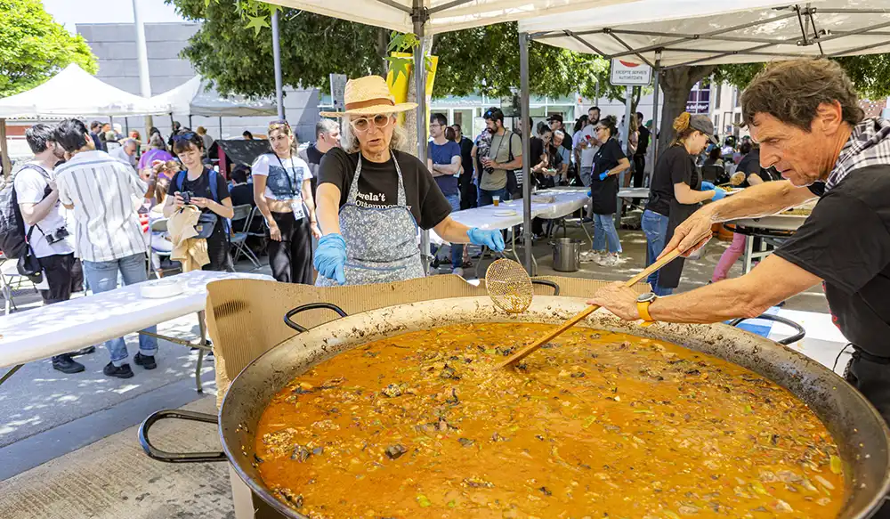 Paella Revela't 2025 - Revela't 2026 People at an outdoor festival cook and serve from a huge pot of orange stew as a volunteer stirs with a skimmer.
