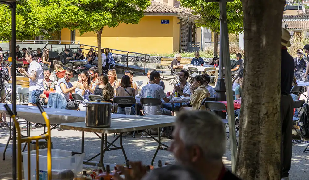 Paella Revela't 2025 - Revela't 2026 Crowd of people dining at outdoor tables in a sunny plaza with a yellow building in the background.