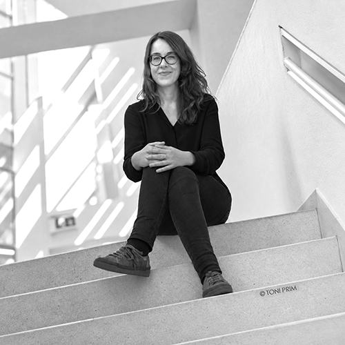 Woman with glasses sits on concrete stairs in a modern interior, hands folded, smiling at the camera in grayscale.