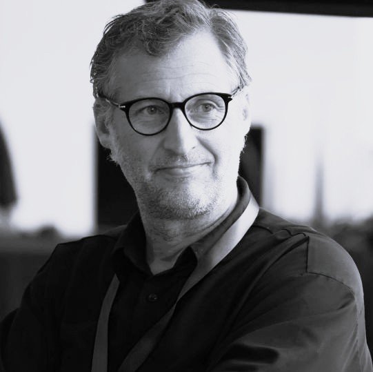 Portrait of a middle-aged man with gray hair and round glasses, looking to the right, wearing a dark shirt in an indoor setting.